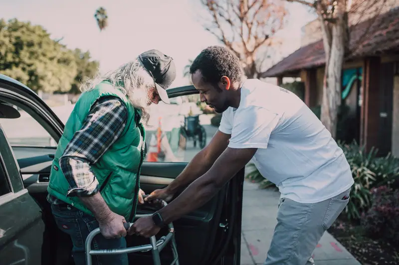 A younger man helping an older man with a walker beside a car